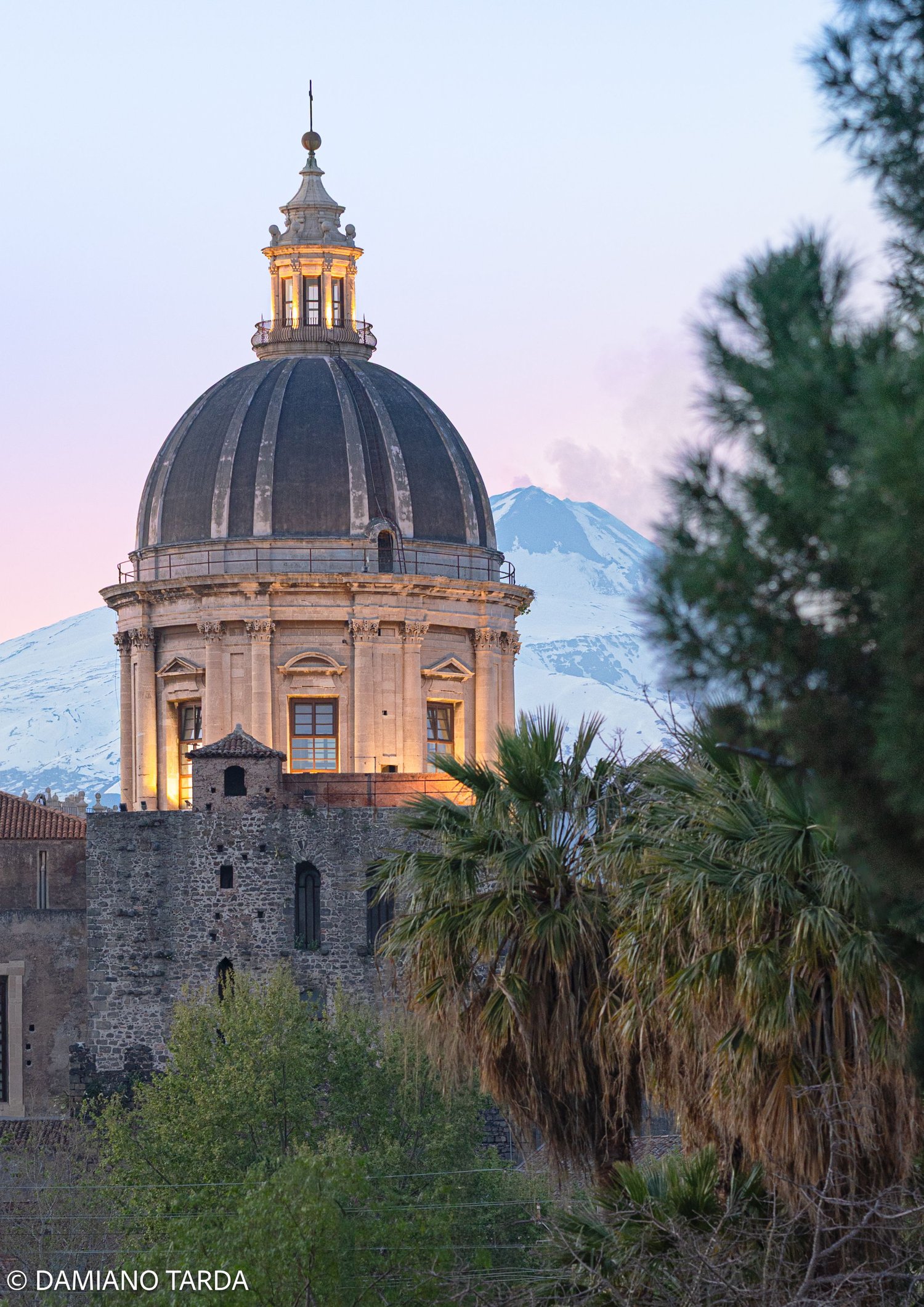 Cupola di una chiesa barocca di Catania al tramonto, con l'Etna innevato sullo sfondo.